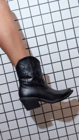 Botas texanas de caña corta, color negro, con taco cuadrado y punta ligeramente afinada. Presentan costuras decorativas en relieve en la caña y cierre lateral interno.