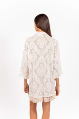 Vestido camisero corto de encaje blanco con diseño ornamental, escote en V con cordón ajustable y ojales metálicos, mangas 3/4 acampanadas y ruedo con detalle de flecos de caracoles tejidos a mano.