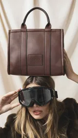 Cartera estructurada de símil cuero marrón, con asas de mano y parche frontal con el logo grabado. Cuenta con compartimento principal y bolsillos internos para mayor organización.