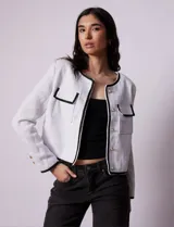 Chaqueta de tweed blanca con ribete negro, escote a la base, abertura central con botones, bolsillos delanteros y puños con aplique de botones perlados.