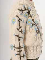 Cárdigan tejido a mano color crudo, con diseño de trenzas y flores bordadas en azul y marrón. Tiene cuello redondo y cierre frontal con botones de madera.