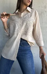 Camisa beige de corte oversize, con cuello clásico y cierre frontal de botones. Presenta un diseño bordado detallado en la parte superior delantera, con mangas largas y un estilo versátil.