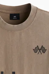 Remera de algodón de punto pesado con efecto lavado, color beige/tostado. Presenta un corte boxy (holgado) con cuello redondo ribeteado y bajo recto. Estampado frontal en negro con texto "Club Issue" y gráficos de banderas a cuadros.