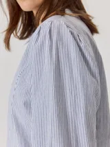 Camisa de denim con silueta levemente entallada, cuello bebé y mangas largas con sutil frunce en los hombros. Presenta costuras curvas decorativas con pespuntes en contraste y cierre frontal con botones.