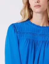 Blusa azul de gasa con pliegues en la parte delantera y detalles de encaje calado. Cuello alto fruncido y mangas largas con puños elastizados. Espalda lisa con abertura tipo gota y botón.