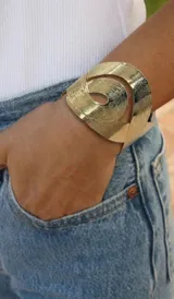Brazalete rígido de acero dorado con diseño abierto y texturizado en líneas concéntricas.