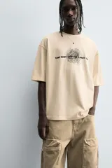 Camiseta de algodón de corte relajado, color beige claro, con cuello redondo y manga corta. Presenta un estampado central de garabatos negros con texto en mayúsculas que dice "TAKE RIGHT STEP FIND A RIGHT CORD".