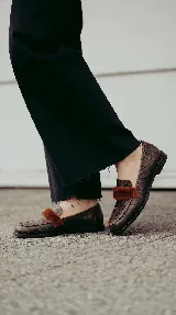Mocasín de cuero con textura gastada, color marrón, con detalle de pelo en la parte superior y suela plana.