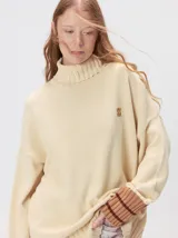 Pullover de punto color rojo, con cuello tortuga, corte holgado y mangas largas con puños acanalados en contraste con rayas blancas. Presenta un logo bordado en el pecho.