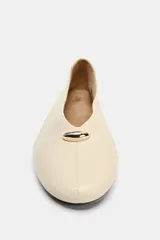 Zapato plano tipo bailarina en piel color beige, con efecto soft y pespuntes en la parte frontal. Presenta escote en pico con detalle metálico y acabado en punta redonda.