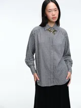 Camisa de corte clásico con estampado de cuadros vichy en tonos azul y blanco. Presenta cuello camisero, cierre frontal con botones ocultos y mangas largas con puños abotonados. Incluye un detalle de broche metálico dorado en el cuello.