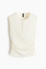 Musculosa de punto acanalado color blanco, ajustada al cuerpo, con escote redondo, tapeta de botones frontal y fruncidos laterales en la cintura.