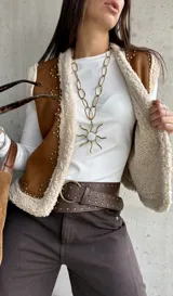 Chaleco de gamuza color camel con interior de corderito sintético, diseño sin mangas, borde frontal con ribete de corderito y detalle de tachas metálicas decorativas.