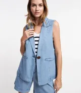 Chaleco largo de jean celeste con escote en pico y cierre con un botón marrón.