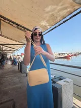 Bandolera pequeña de diseño rectangular en color beige, confeccionada en cuero sintético. Presenta una correa larga ajustable del mismo material y cierre superior con cremallera. Incluye el logo de la marca grabado en la parte frontal.