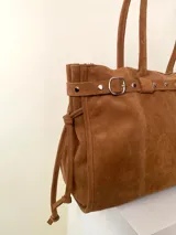 Bolso grande de cuero marrón con textura tipo cocodrilo, estructura rígida y asas para llevar al hombro. Tiene una tira con tachas y hebillas en cada extremo, también lleva lazos en los costados.