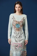 Vestido largo de corte recto y manga larga, con cuello redondo. Presenta un estampado artístico integral con motivos botánicos, paisajes montañosos y fauna, en tonos celestes, marrones y verdes.