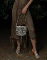 Cartera compacta de cuero con estampado animal print tipo dálmata y solapa frontal. Cuenta con asa trenzada, clip de cierre, costuras visibles en los bordes y detalle de flecos colgantes.