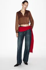 Camisa corta de tejido satinado color marrón, con cuello solapa, escote en pico y manga larga. Presenta cierre frontal con botones forrados a tono.