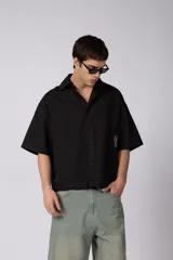 Camisa de manga corta negra 100% algodón, con calce boxy, cuello camisero, cierre frontal con botones y detalle de bordado blanco en la parte inferior.