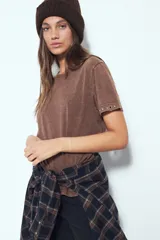 Remera de manga corta color marrón con escote a la base y calce relajado. Las mangas presentan terminación con ojales metálicos.