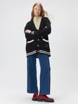 Cardigan de punto trenzado en color negro con detalles en color crema en los bolsillos, puños y ruedo. Presenta un diseño oversize, cierre frontal con botones y bordado decorativo en uno de los bolsillos delanteros.