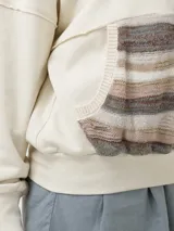 Buzo de cuello alto con medio cierre frontal, confeccionado en algodón orgánico. Presenta un bolsillo canguro delantero con un diseño tejido artesanal en tonos tierra y texturas variadas.