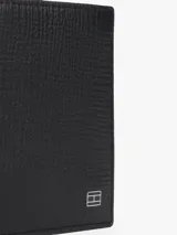 Billetera plegable de piel color negro con textura, diseño compacto y detalle de placa esmaltada con el logo de Tommy Hilfiger en la esquina inferior.