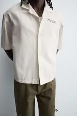 Camisa de corte relajado confeccionada en algodón, con cuello bowling y manga corta. Presenta cierre frontal con botones y detalle de texto estampado en el pecho.