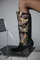 Bota de caña alta confeccionada en gamuza negra, con un diseño artesanal de parches en cueros texturizados y metalizados con motivos florales. Presenta un taco cónico y punta almendrada.