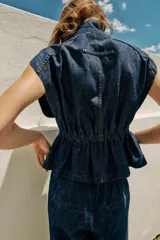 Chaleco de denim azul oscuro con cuello alto, manga sisa y cintura ajustable con elástico. Cierre frontal con botones a presión metálicos.