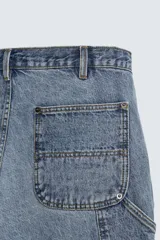 Pantalón de jean estilo carpintero con corte relaxed fit, tiro medio y pierna recta. Presenta un lavado azul medio con efecto desgastado y roturas en las rodillas. Incluye bolsillos laterales tipo plastrón, tira multifunción en la pierna, cierre frontal con cremallera y botón.