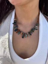 Collar corto de diseño artesanal compuesto por bastones de piedra natural en tonos verdes, marrones y grises, ensartados junto a pequeñas mostacillas ámbar en la parte posterior.