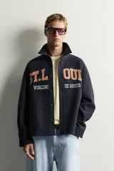 Sobrecamisa de ajuste relajado en color azul marino, con cuello de solapa, manga larga con puño abotonado y cierre frontal con botones. Presenta bordados contrastantes en el frente con la inscripción "St. Louis Worldwide Goods" en tonos naranja y blanco.