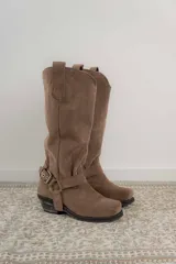 Bota estilo tejana de caña alta, confeccionada en cuero marrón con acabado texturizado. Presenta punta cuadrada, taco bajo y una correa con hebilla metálica ajustable y estribo desmontable en la zona del tobillo.