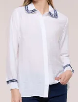 Camisa de crepe liviana y texturada color blanco, de silueta relajada y holgada, con cuello camisero clásico y puños con detalles de bordado de pasamanería con terminación deshilachada y aplique de perlas. Cuenta con cartera de botones oculta al frente y mangas largas.