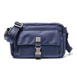 Bandolera de diseño rectangular en color azul, confeccionada en material textil. Presenta un compartimento frontal con solapa, cierre de hebilla metálica a presión y detalle de placa metálica cuadrada. Incluye correa ajustable con logo grabado y herrajes metálicos.