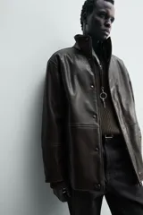 Cazadora de corte relajado confeccionada en tejido efecto piel color marrón oscuro. Presenta cuello subido con interior forrado en tejido de cuadros, manga larga, bolsillos de vivo en la cadera y cierre frontal con cremallera oculta por solapa con botones a presión.
