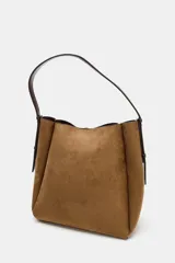 Bolso tipo saca de gran tamaño confeccionado en material efecto gamuza color verde oliva. Presenta un diseño estructurado con pespuntes decorativos, una anilla metálica dorada en el frente y cierre mediante botón imantado. Incluye un asa de hombro ajustable y bolsillo interior con cremallera.