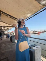 Bandolera pequeña de diseño rectangular en color beige, confeccionada en cuero sintético. Presenta una correa larga ajustable del mismo material y cierre superior con cremallera. Incluye el logo de la marca grabado en la parte frontal.