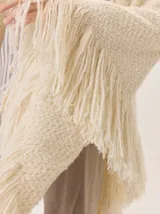 Triángulo de lana color beige con maxi flecos, diseñado por Margara Shaw en colaboración con Manos del Uruguay. Confeccionado con una mezcla de lana merino, mohair, viscosa, algodón y otras fibras, incluyendo lurex y un hilado teñido a mano con lentejuelas.