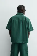 Camisa de denim de algodón color verde oscuro, de corte relajado (relaxed fit) y manga corta. Posee cuello solapa, bolsillo plastrón en el pecho con bordado a tono y cierre frontal de botones.