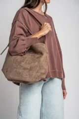Cartera tipo shopper de gamuza sintética color marrón, con diseño estructurado, asa de hombro ajustable y detalle de placa metálica dorada con logo en la parte frontal.