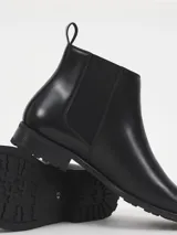 Botin tipo Chelsea de cuero sintético negro, con paneles laterales elásticos, tirador trasero y suela de goma dentada.