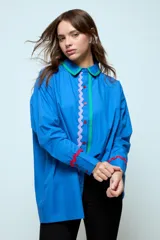 Camisa azul de corte oversize, con cierre frontal de botones y detalles de cintas onduladas en colores contrastantes a lo largo de la botonadura y en los puños. Presenta cuello clásico y mangas largas.