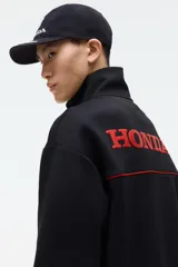 Campera deportiva negra de efecto neopreno con cierre bidireccional, cuello elevado y bolsillos laterales. Presenta detalles de ribetes rojos en el pecho y el logo de Honda bordado en rojo.