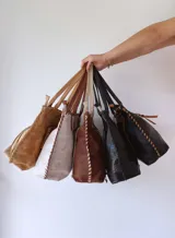 Cartera tipo tote bag de gamuza negra, con detalles de costuras decorativas en forma de X y tiras de cuero entrelazadas en el frente. Cuenta con doble asa de hombro y cierre superior.