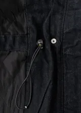 Parka de denim negro de corte largo, con cuello alto, cierre frontal oculto y múltiples bolsillos con solapa en el pecho y los costados. Cuenta con cordón ajustable en la cintura.