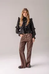 Camisa negra de corte entallado con volados decorativos en el cuello, la botonadura frontal, los hombros y las mangas. Presenta mangas largas con puños ajustados y un ruedo con volado inferior.