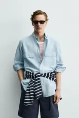 Camisa de corte relajado color celeste, confeccionada en mezcla de algodón y seda. Presenta cuello solapa, cierre frontal de botones, manga larga con puños abotonados y dos bolsillos de plastrón con solapa en el pecho.